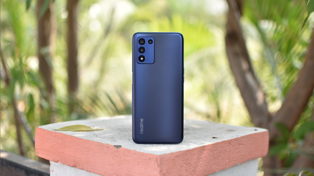  A Realme 12 Pro 5G smartphone in dark blue is placed on a concrete surface outdoors, leaning against a small block with a leaf on it.